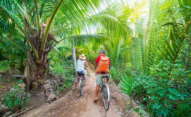 Pareja de turistas montando bicicleta en la región del Delta del Mekong, Ben Tre, Vietnam del Sur. La mujer y el hombre se divierten en bicicleta en el sendero entre bosques tropicales verdes y palmeras de coco. Vista trasera.