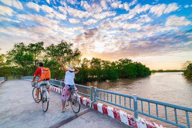 Pareja de turistas montando bicicleta en la región del Delta del Mekong, Ben Tre, Vietnam del Sur. Mujer y hombre divirtiéndose ciclismo entre bosques tropicales verdes y canales de agua. Vista trasera puesta de sol cielo dramático.