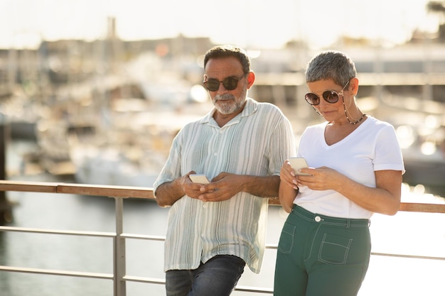 Pareja usando teléfonos inteligentes planeando vacaciones con aplicaciones de viaje en el puerto deportivo