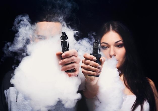 Pareja vaping. Un joven irreconocible y una mujer en las nubes de humo mostrando sus vaporizadores a la cámara en el fondo negro del estudio. Concepto de relación y adicción a vape con espacio de copia