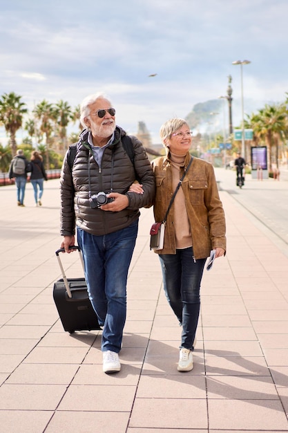 Una pareja vertical de turistas caucásicos mayores felices paseando abrazados sin preocupaciones con su equipaje.