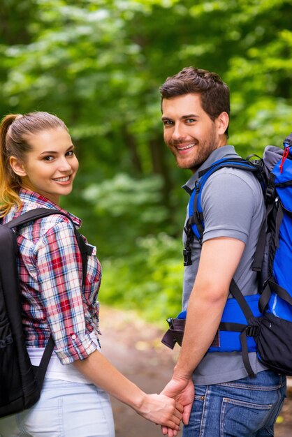 Pareja viajando con mochilas. Feliz pareja amorosa joven llevando mochilas y mirando por encima del hombro con una sonrisa mientras camina por el sendero del bosque