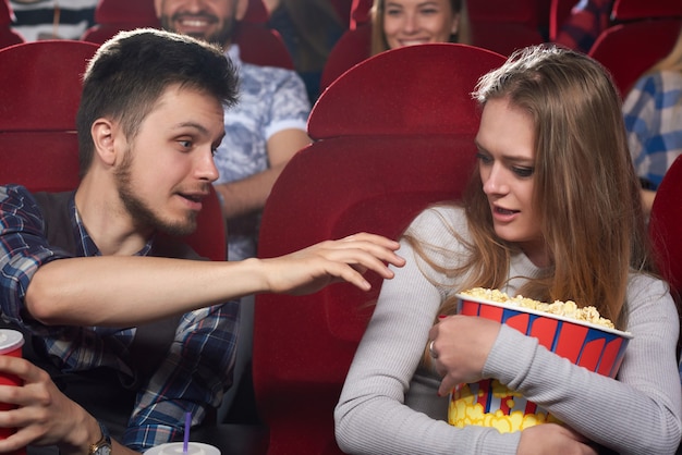 Pareja viendo películas en el cine, novio enojado tirando de la mano a las palomitas de maíz y su novia codiciosa abrazando un gran cubo y gritando.