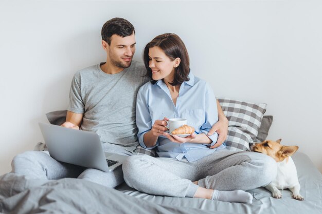 Foto pareja viendo video en la computadora portátil mientras está sentada con el perro en la cama contra la pared en casa