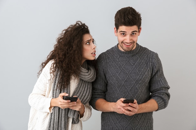 Pareja vistiendo suéteres que se encuentran aisladas sobre la pared gris, utilizando teléfonos móviles, mujer molesta mirando a un hombre feliz