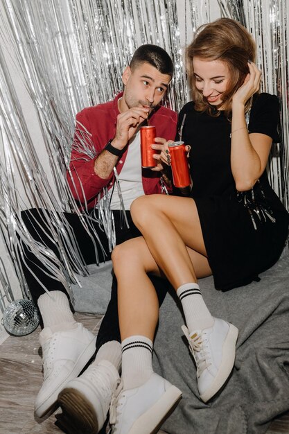 pareja de zapatillas sentada en el suelo bebiendo refrescos y divirtiéndose contra la cortina de plata