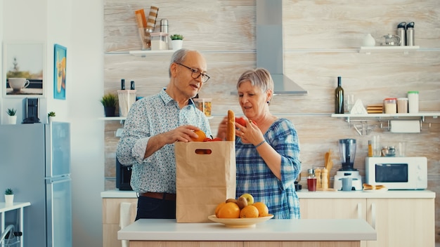 Las parejas ancianas que llegan del supermercado con la bolsa de la compra y desembalaje en la cocina. Personas mayores jubiladas disfrutando de la vida, pasando tiempo ayudándose unos a otros.