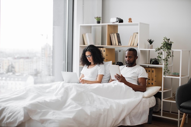 Parejas románticas descansando en la cama con dispositivos electrónicos