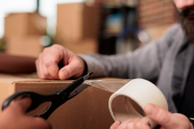 Parejas de vida casadas que usan cinta adhesiva y tijeras para empacar muebles en cajas, mudándose juntos a la propiedad del hogar. Embalar cosas con rodillo adhesivo para mudarse en piso alquilado. De cerca.