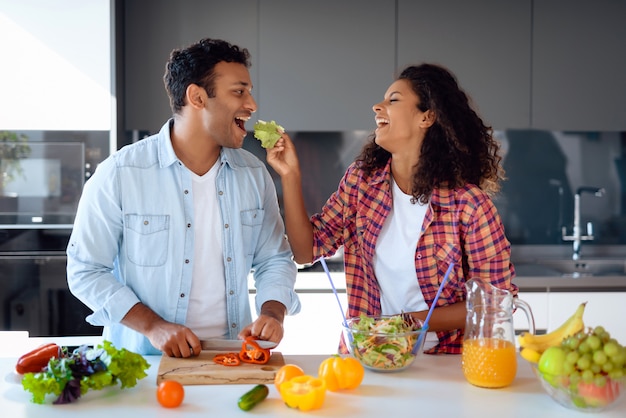 Pares afro-americanos que cozinham a salada na cozinha.