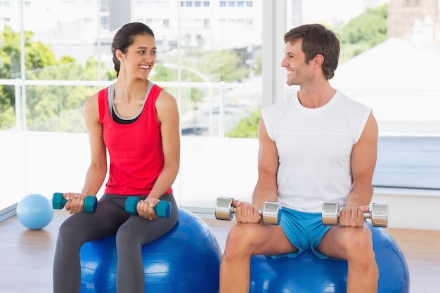 Pares felices que levanta pesas de gimnasia mientras que se sienta en bolas de la aptitud en gimnasio