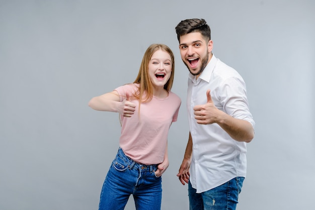 Pares jovenes felices del hombre y de la mujer que muestran los pulgares para arriba aislados sobre fondo gris