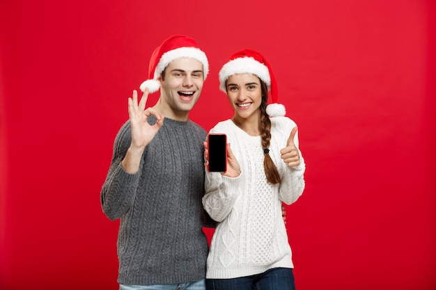 Pares jovenes felices en suéteres de la Navidad que muestran gesto aceptable con el teléfono móvil.