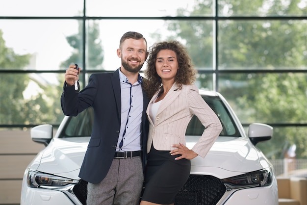 Foto pares sonrientes que se colocan cerca del coche blanco, mirando la cámara.