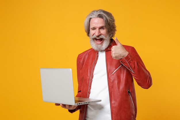 Parpadeando a un anciano bigote de pelo gris con barba y chaqueta de cuero rojo posando aislado en un fondo amarillo. Concepto de estilo de vida de las personas. Simulacros de espacio de copia. Sostenga la computadora portátil, mostrando el pulgar hacia arriba.