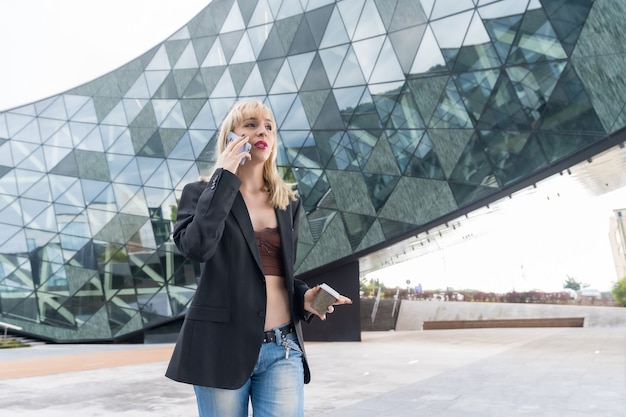 Parque empresarial chica emprendedora en un edificio moderno con vidrio coloreado, joven rubia sonriente en traje haciendo una llamada de trabajo