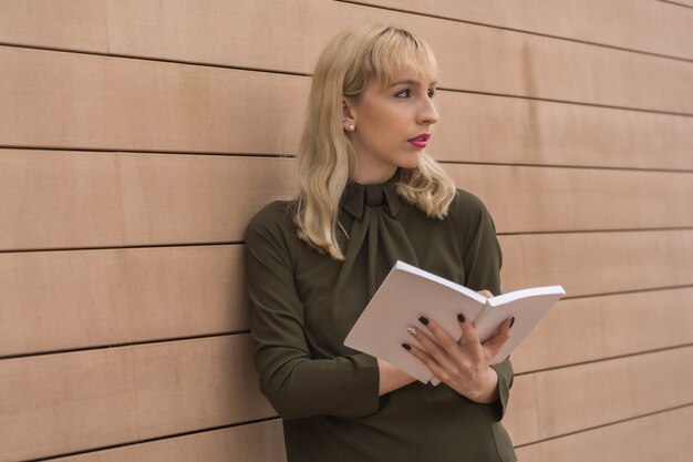 Parque empresarial empresarial chica apuntando notas en el diario, joven rubia con una camisa verde apoyada contra una pared de madera, mirando a la derecha