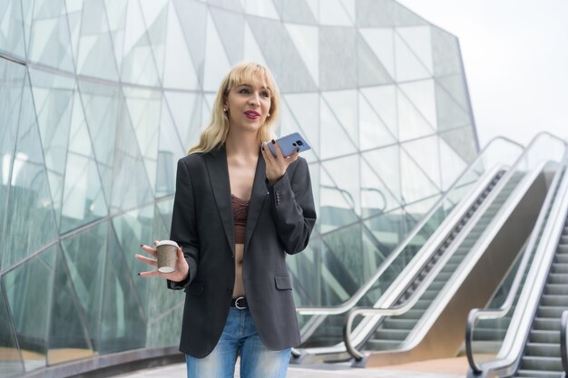 Parque empresarial feminino empreendedor em um edifício moderno com escada rolante, jovem loira de terno enviando uma nota de voz com um café para viagem