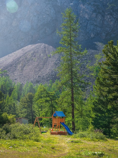 Parque infantil bajo un alto alerce en el fondo de verdes montañas boscosas. Vacaciones familiares ecológicas en el concepto de montañas y bosques. Vista vertical.