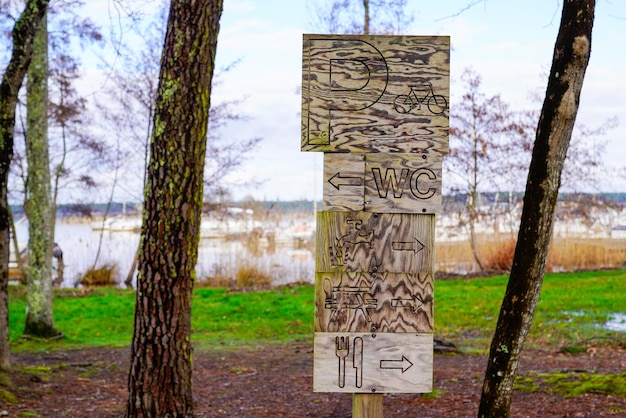 Parque de madera con varias señales de flecha que incluyen wc y estacionamiento de bicicletas Baños y punto de agua para picnic a un lado del lago de agua del río