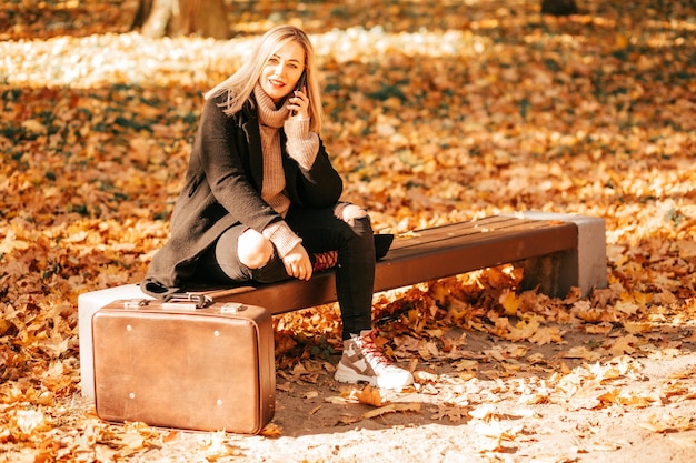 En el parque de otoño de la ciudad, entre hojas doradas caídas, una atractiva rubia con abrigo negro se sienta en un banco y habla por teléfono inteligente, la vieja maleta está parada a su lado. Estilo de vida. La vida en el momento