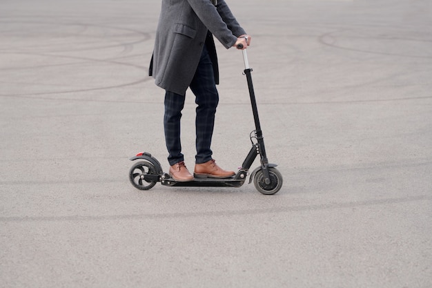 Parte baixa de um jovem empresário contemporâneo de casaco, calça e botas em pé na scooter elétrica enquanto se dirige para o trabalho pela manhã