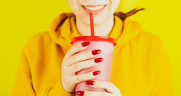 Parte del cuerpo de la mujer con manicura roja sosteniendo una taza grande Persona irreconocible de cultivo bebiendo a través de paja de refresco sobre fondo amarillo