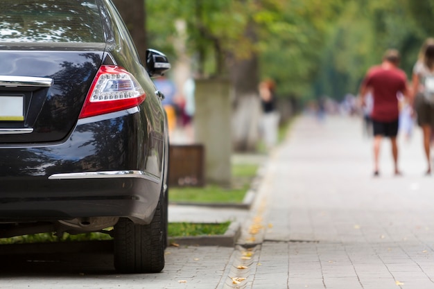 Parte da vista traseira do carro brilhante de luxo preto estacionado no pavimento da zona pedonal da cidade no fundo das silhuetas borradas de pessoas caminhando ao longo do beco verde ensolarado de verão. conceito de estilo de vida moderno.