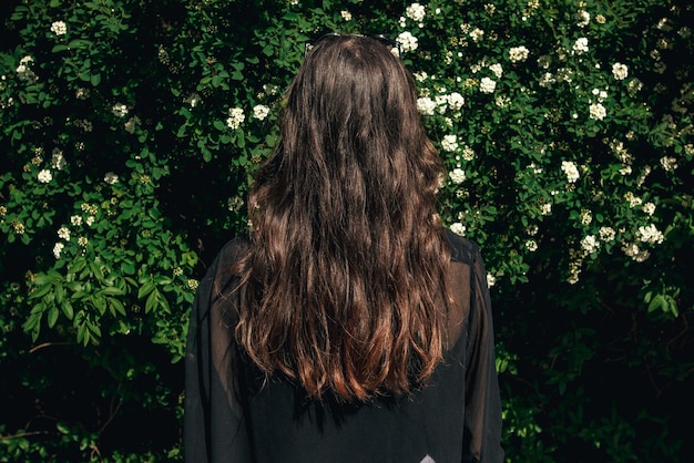 Parte de trás da linda menina elegante Hipster com cabelo bonito em arbustos florais em dia ensolarado Boho Woman com óculos de sol em roupa elegante, aproveitando o dia no momento calmo do jardim