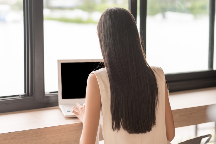 Parte de trás da mulher sentada e usando o computador portátil ao lado das janelas