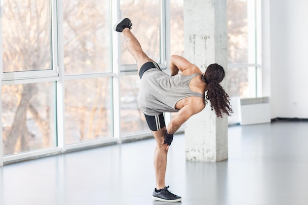 Parte de trás do homem adulto jovem musculoso saudável com cabelo comprido encaracolado está posando na posição profissional de kickboxing com as pernas levantadas, mostrando a técnica de combate Fundo da janela interna artes marciais