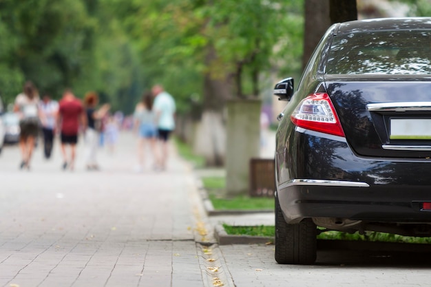 Parte de vista traseira do carro brilhante de luxo preto estacionado no pavimento da zona pedonal da cidade no fundo das silhuetas borradas de pessoas caminhando ao longo do beco verde ensolarado de verão. Conceito de estilo de vida moderno.