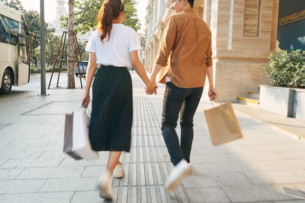 La parte inferior del cuerpo de una joven pareja de turistas caminando por los escaparates de las tiendas y sosteniendo bolsas de papel en una ciudad de destino.