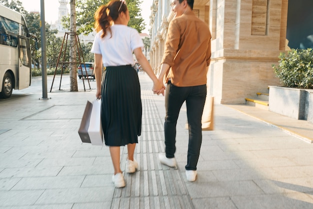 La parte inferior del cuerpo de una joven pareja de turistas caminando por los escaparates de las tiendas y sosteniendo bolsas de papel en una ciudad de destino.