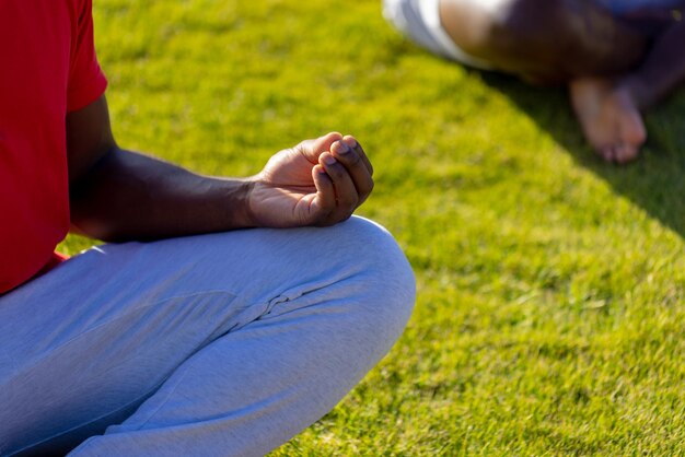 Foto parte intermediária do homem afro-americano sentado na grama, fazendo ioga e meditando no jardim. esporte, estilo de vida saudável e ativo, verão, sol, inalterado.