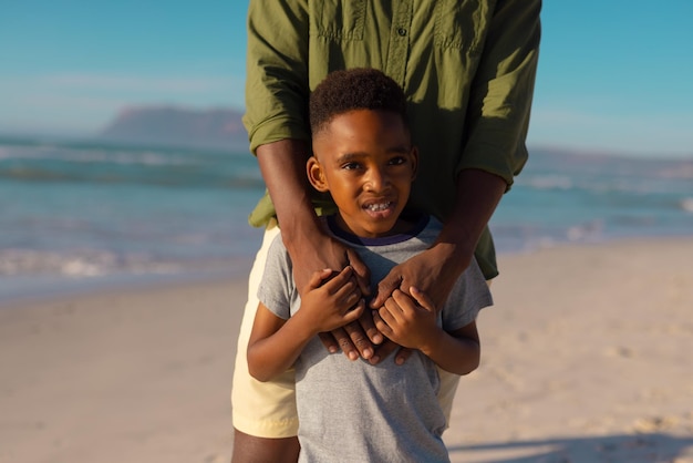 Parte intermediária do jovem afro-americano segurando as mãos do filho em pé contra o mar e o céu