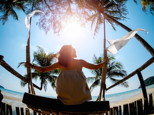 La parte posterior de una mujer asiática vestida de forma informal se sienta en un columpio frente a la playa en verano con la luz del sol Mujer solitaria relajándose en el mar y mirando al cielo Vista del paisaje marino Vacaciones Viajes vacaciones