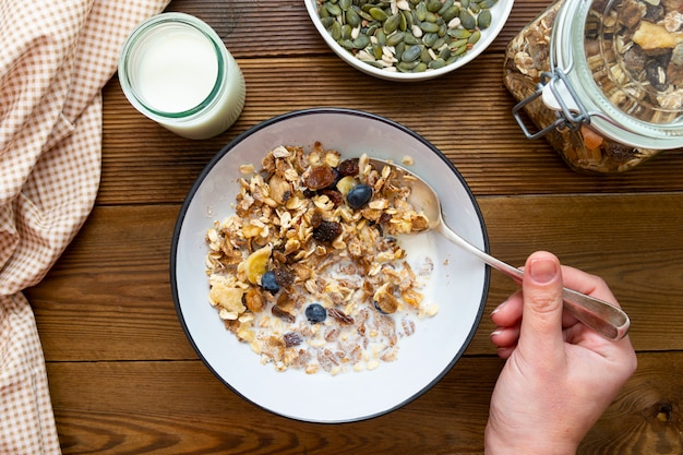 Parte superior de la mano sosteniendo una cuchara de avena para el desayuno, granola con frutos secos y arándanos, leche y miel.