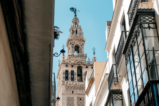 Parte superior de la torre gótica que se asoma entre edificios en una calle estrecha en un día soleado