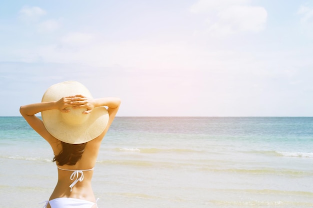 Parte traseira jovem pele bronzeada em chapéu de sol e biquíni em pé com os braços levantados para a cabeça curtindo a viagem de férias na praia e aproveitar a vida no mar, olhando a vista do oceano da praia em um dia quente de verão