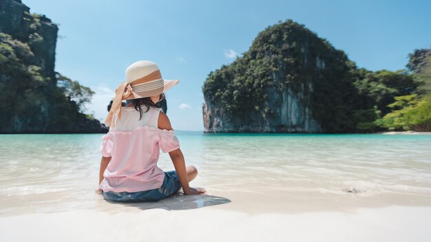 Parte trasera de la niña asiática con sombrero sentado en la playa.