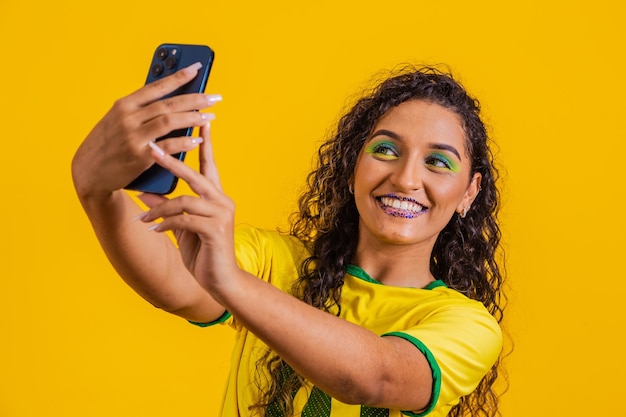 Partidario de Brasil Fan de la mujer brasileña celebrando el partido de fútbol con fondo amarillo Brasil colorea el teléfono inteligente Selfie