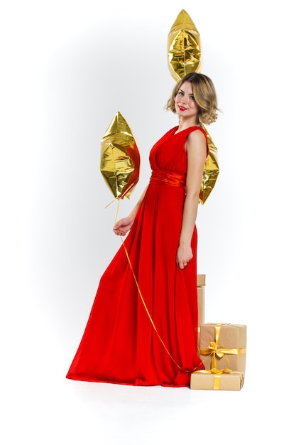 Partyfoto der sexy Dame der Eleganz im roten Kleid mit den roten Lippen und dem blonden schönen lockigen Haar, lächelnd. Hintergrund von Goldluftballons und Geschenken. Konzept des Verkaufs