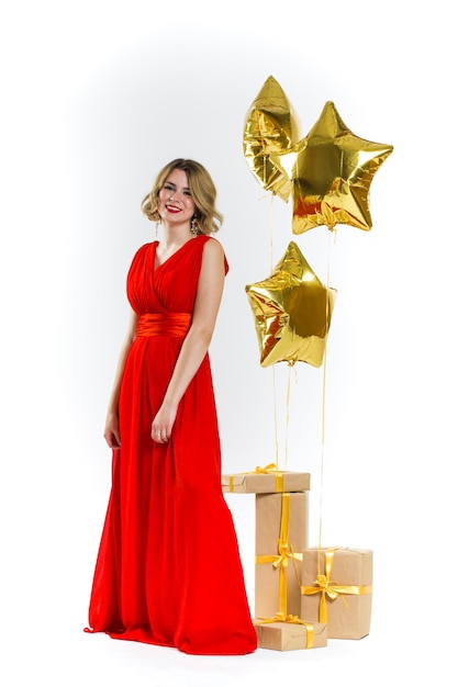 Partyfoto der sexy Dame der Eleganz im roten Kleid mit den roten Lippen und dem blonden schönen lockigen Haar, lächelnd. Hintergrund von Goldluftballons und Geschenken. Konzept des Verkaufs