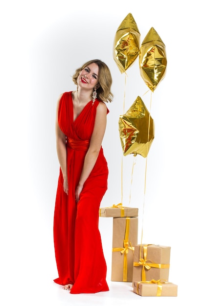 Partyfoto der sexy Dame der Eleganz im roten Kleid mit den roten Lippen und dem blonden schönen lockigen Haar, lächelnd. Hintergrund von Goldluftballons und Geschenken. Konzept des Verkaufs