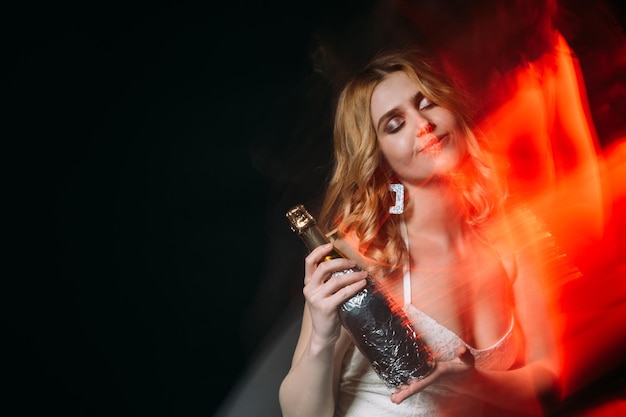 Partyfreude Inspirierte Frau Urlaubsgetränk Festliche Stimmung Fröhliche, verträumte Dame im weißen Kleid, die die Augen hält und eine Champagnerflasche mit langer Belichtungszeit defokussiert, rotes Licht isoliert, schwarzer Kopierraum hält