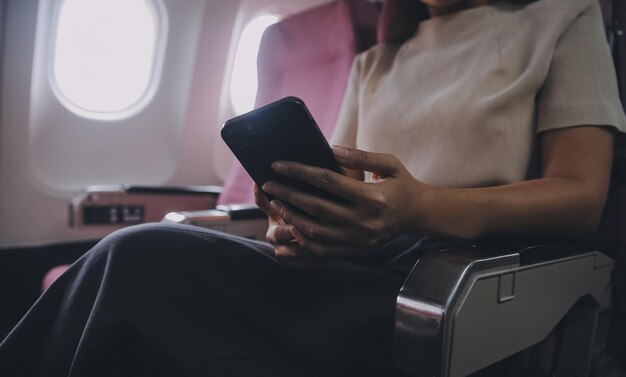 Pasajera asiática sentada en un avión cerca de una ventana y leyendo noticias de las redes sociales o usando aplicaciones de viaje en un teléfono inteligente