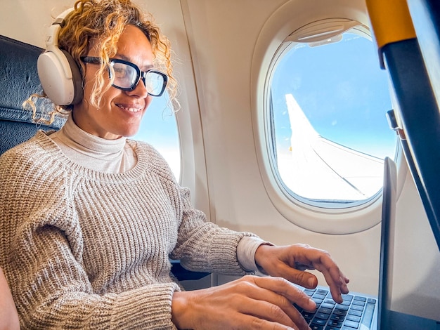 Foto pasajera rubia atractiva con cabello rizado de un avión sentada en un asiento cómodo escuchando música en auriculares mientras trabaja en una computadora portátil moderna usando conexión inalámbrica a bordo