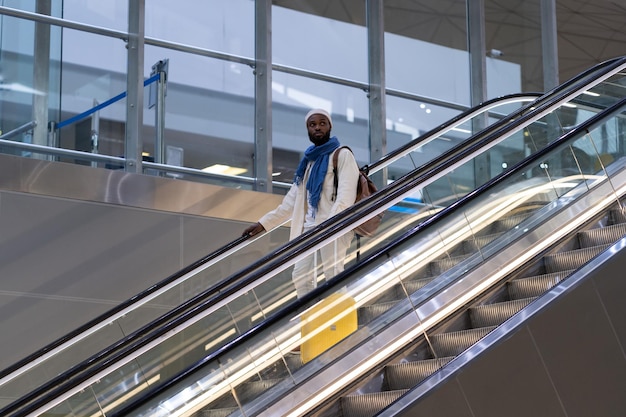 El pasajero afroamericano con maleta se para en la escalera mecánica sostiene el pasamanos en la terminal del aeropuerto