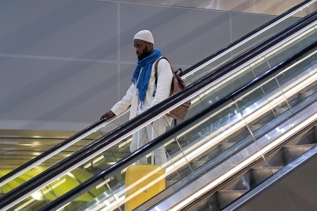 El pasajero afroamericano con maleta se para en la escalera mecánica sostiene el pasamanos en la terminal del aeropuerto